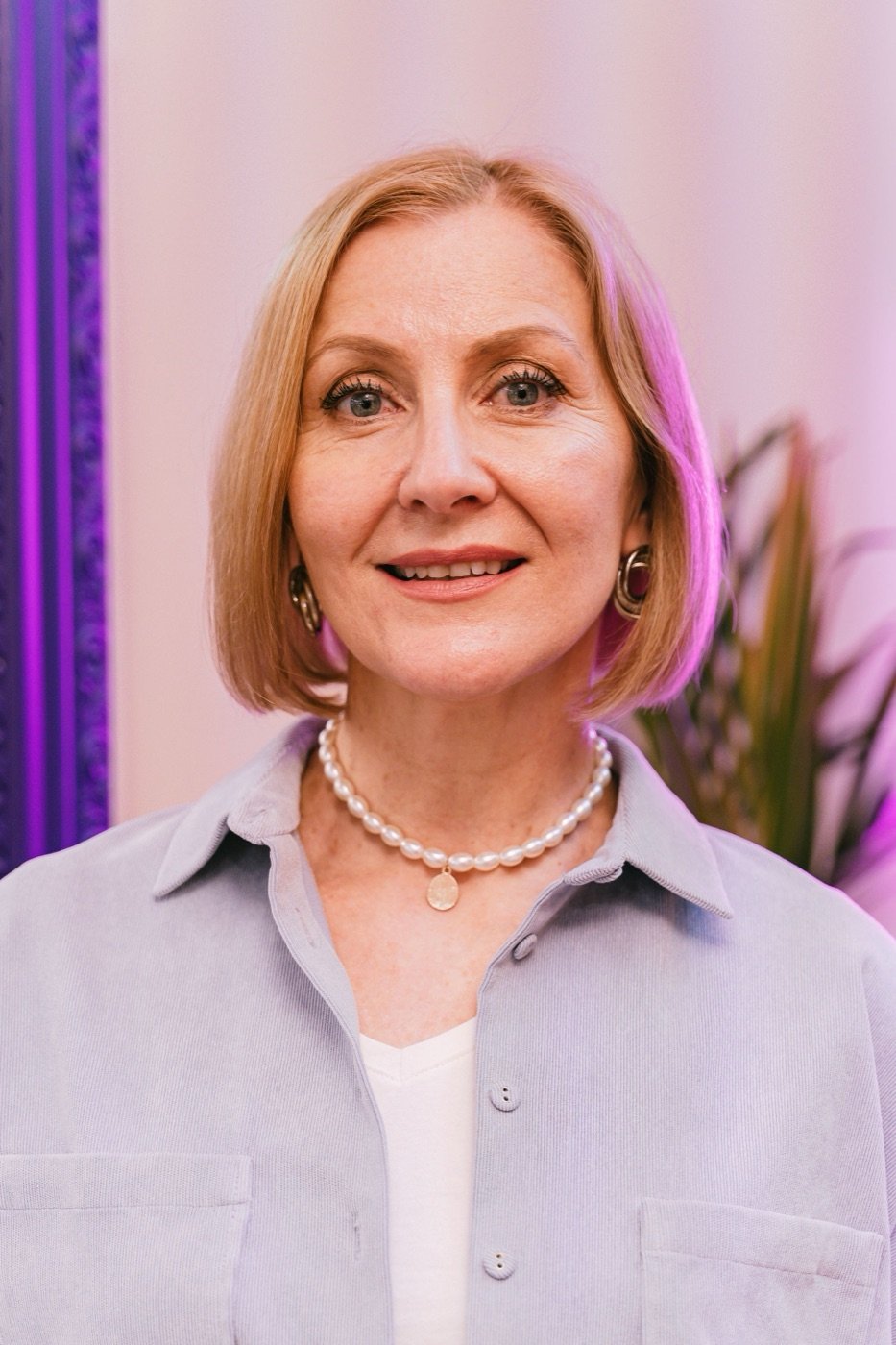 A woman with short blonde hair and pearls, in lilac light