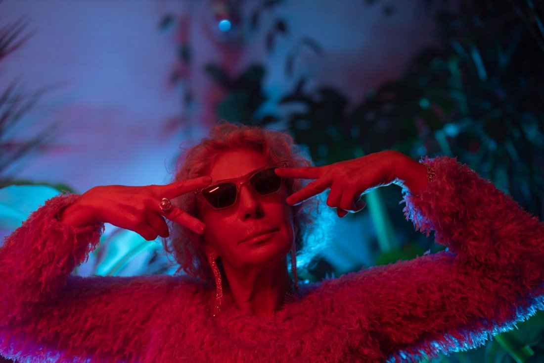 A woman in pink fur and sunglasses, in red and teal light